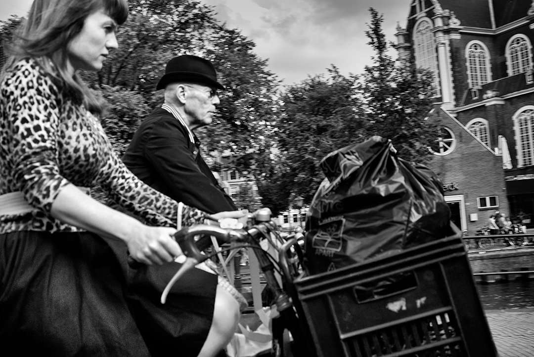 A woman on a bicycle passes a man in a bowler hat and a covered carriage.